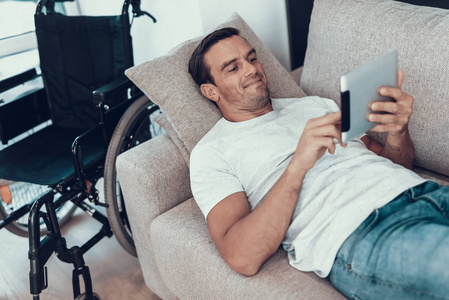 坐在沙发上休息的男人，坐在轮椅旁的写字板上。开朗英俊微笑的年轻残疾人躺在现代公寓舒适明亮的沙发上，用垫子上网