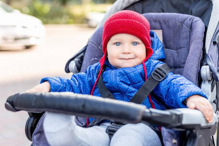 可爱的小男孩坐在婴儿车里，在寒冷的秋天或冬天散步时微笑。可爱的孩子穿着蓝色夹克和针织红色帽子在户外。快乐健康的童年理念..
