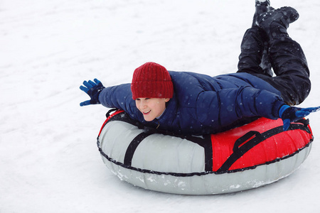 穿着蓝色夹克和红色帽子的孩子在雪管上玩得很开心。 男孩在骑油管。 冬季娱乐活动。 积极的运动生活方式