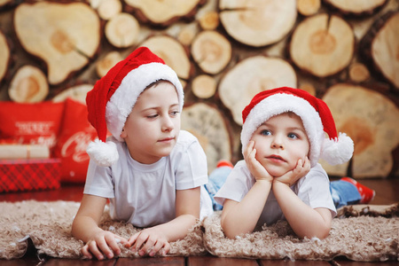 两个戴着圣诞帽的兄弟男孩躺在新年树附近的地毯上，在砍树的背景下笑着。