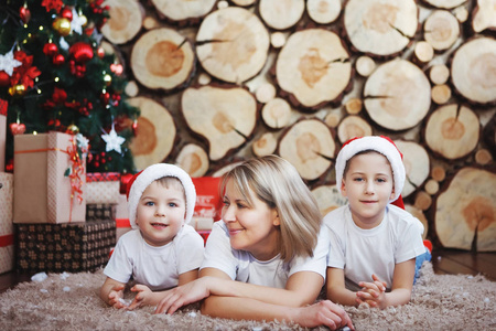母亲带着两个儿子戴着圣诞帽，躺在新年树附近，背景是砍树。