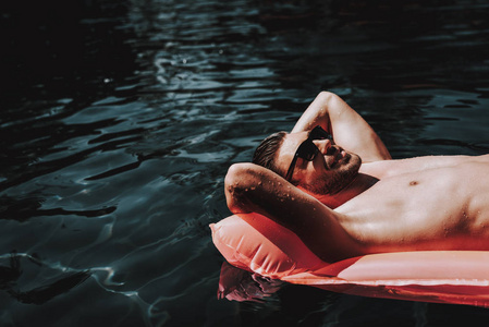 年轻的微笑男人戴着太阳镜躺在气垫上。 英俊的胡须男，戴着太阳镜，躺在户外游泳池的充气床垫上放松。 暑假概念