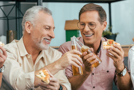快乐的成熟男人在家吃披萨和叮叮作响的啤酒瓶