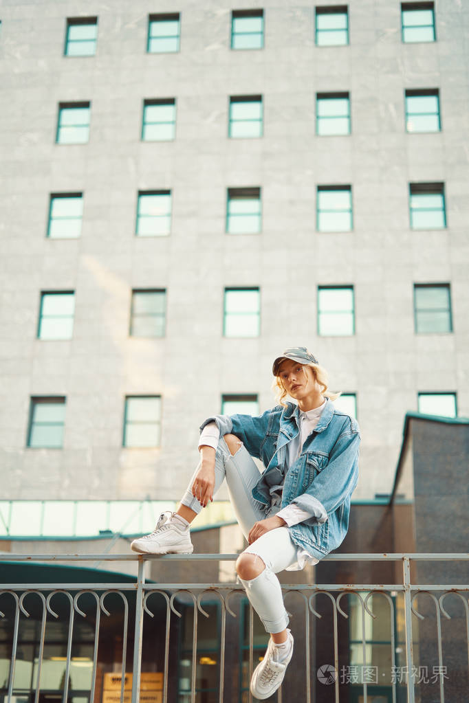 美丽的女孩坐在欧洲城市的栅栏栏杆上背景是现代建筑同时观看城市生活