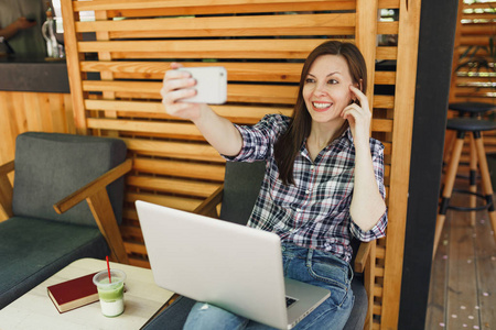 女士在木制户外街道夏季咖啡店，坐在笔记本电脑电脑上，在手机上自拍，在空闲时间放松。 移动办公室。 生活方式自由职业商业概念