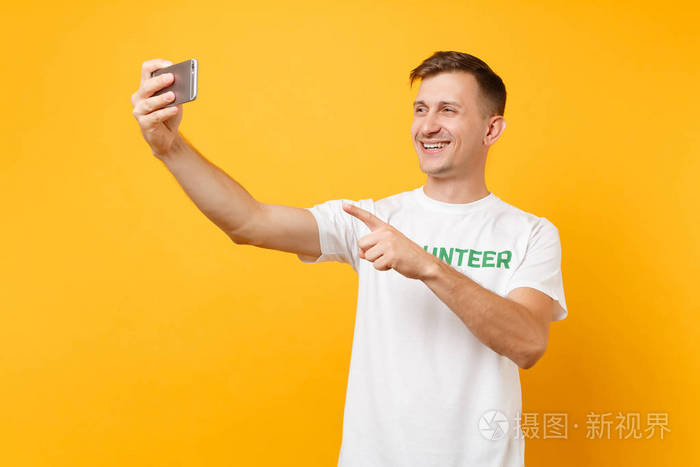 白色T恤男子肖像，文字铭文，绿色标题，志愿者，在手机上自拍，孤立在黄色背景上。 自愿无偿援助慈善恩典工作概念