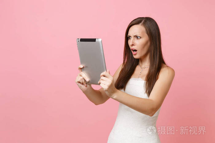 震惊困惑的新娘女人穿着白色婚纱在平板电脑电脑上工作,在粉彩背景上