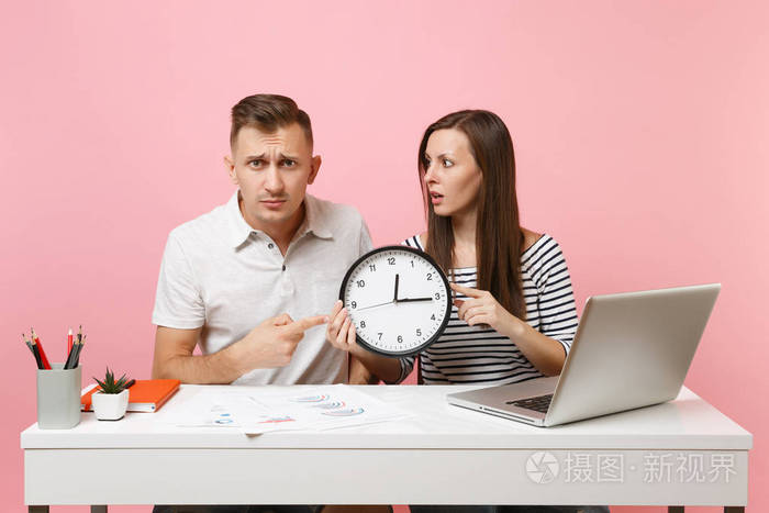 两个年轻的乐趣震惊商务女性男性同事坐在白色办公桌上工作,时钟