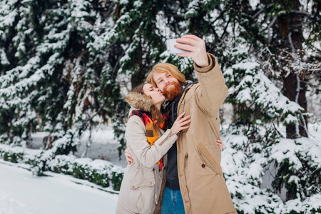 这家伙把智能手机放在拉长的手上,在冬天,自拍照片女孩亲吻她的脸颊.
