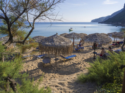 沙和鹅卵石海滩和绿松石海岸在AgiosGeorgiosPagon在科孚岛希腊，日光浴的人民和伞，在波尔图蒂莫尼湾克莱尔蓝天背。