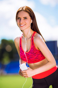 一位身穿运动服的年轻运动微笑女子的肖像，她在体育场上做运动，用耳机听音乐