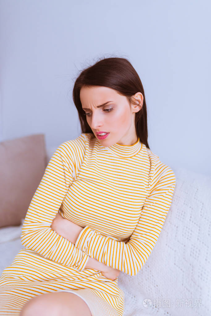 迷人的,忧心忡忡的黑发女人坐在沙发上,把胳膊放在房间里的肚子上