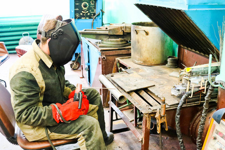 一名男性工人在一个维修设施的冶金工厂的车间里, 在一个焊接站的金属管道上焊接一个保护面罩的焊工。
