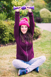 年轻的运动型微笑的女人坐在公园里的莲花姿势里，手里拿着哑铃做运动，把手举起来