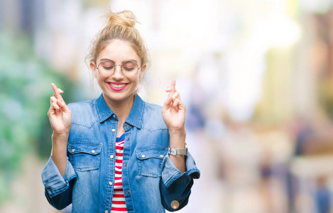 年轻漂亮的金发女人戴着眼镜在孤立的背景上微笑着交叉手指与希望和眼睛闭上。 运气和迷信的概念。