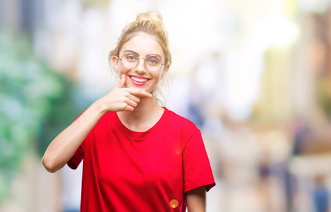 年轻漂亮的金发女人穿着红色的T恤和眼镜，戴着孤立的背景，自信地看着相机，微笑着交叉的手臂和举起的手在下巴上。 积极思考。