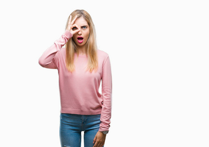 年轻漂亮的金发女人穿着粉红色的冬季毛衣，在孤立的背景上做着OK的手势，惊讶的脸眼睛透过手指看着。 不相信的表情。
