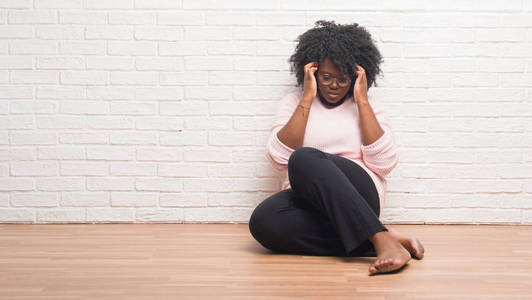 年轻的非裔美国妇女坐在地板上，在家里，手在头上疼痛，因为压力。 偏头痛。