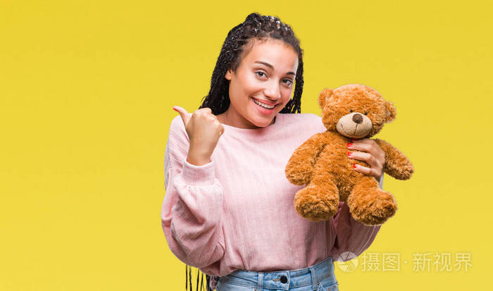 年轻的非裔美国女孩抱着玩具熊在孤立的背景上指指点点，用拇指向旁边露出快乐的脸微笑