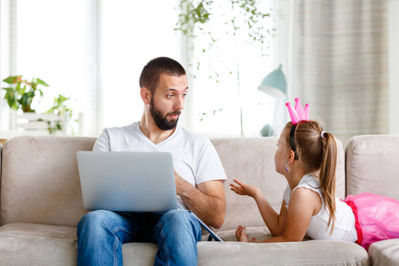 年轻的父亲和女儿花时间在家里，坐在客厅的沙发上说话。 男人使用笔记本电脑和女孩数字平板电脑。