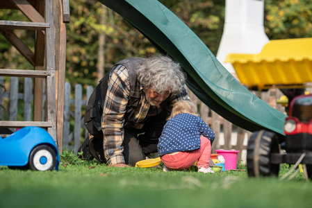 爷爷和他的孙女在后院玩，因为他们坐在草地上在滑梯下玩沙子玩具和水桶。