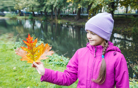 美丽的微笑女孩，戴着蓝帽子，穿着洋红夹克，在秋日户外放着五颜六色的秋叶