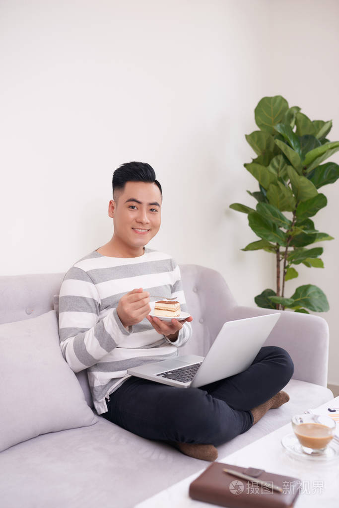 快乐的男人用笔记本电脑工作在沙发上吃蛋糕