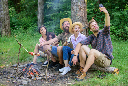 篝火附近的朋友享受度假和烘焙食品。人拍照附近篝火自然背景。游客坐在篝火旁拍摄自拍照片智能手机。朋友度假捕捉时刻