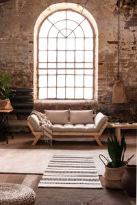 窗户上方灰色木制沙发在宽敞的阁楼内部，瓦比萨比风格，植物和地毯。真实照片