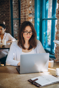 年轻的留胡子的女商人穿着牛仔衬衫坐在办公室的桌子上，正在使用笔记本电脑，屏幕上有图表和图表。 工作的人
