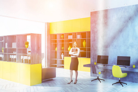 金发女商人在创业办公室，混凝土墙，白色木制地板，黄色和木制书柜，长桌上有电脑显示器。 调色图像