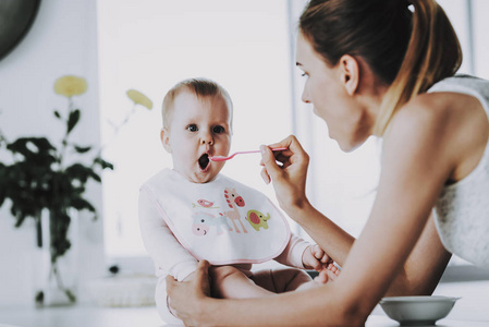 快乐的妈妈在室内用勺子喂可爱的宝宝。 妈妈在明亮的客厅里潜水健康的食物给可爱的饥饿的蹒跚学步的孩子。 幸福的家庭在厨房吃早餐。 