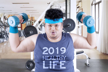 肥胖男子的形象是举起两个杠铃在健身房中心与文字2019年健康生活在他的衬衫上
