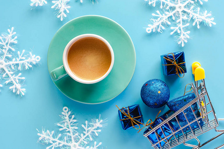 一杯带有圣诞节雪花礼品盒的咖啡，在迷你购物车或蓝色背景的手推车中有闪闪发光的球和靴子装饰，用于圣诞节和假日的饮料和饮料