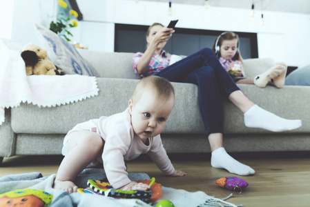 幸福的家庭在明亮的客厅里休息。 婴儿在地板上爬行和玩玩具，而母亲在舒适的沙发上看电视和姐姐用耳机听音乐