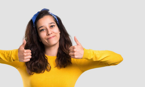 年轻女孩，头上戴着黄色毛衣和蓝色手帕，竖起大拇指，微笑着，因为在孤立的灰色背景上取得了成功
