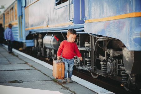 可爱的小男孩穿着红色毛衣在火车站附近的火车上，带着复古的棕色手提箱。 准备好度假了。 站台上的年轻旅行者。