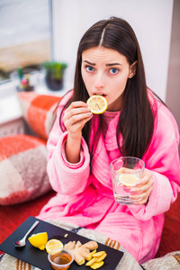 年轻生病的女人坐在家里的床上，拿着一杯水和药品，吃着柠檬的画像