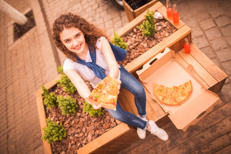 那个年轻漂亮的女人坐在户外，微笑着，手里拿着一块比萨饼