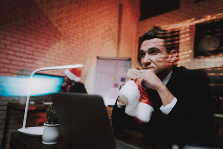 疲惫的年轻商人在新年前夕就职。 办公室里的圣诞树。 笔记本电脑在桌子上。 商业概念。 穿黑色西装的人。 庆祝新年。 使用数字设备