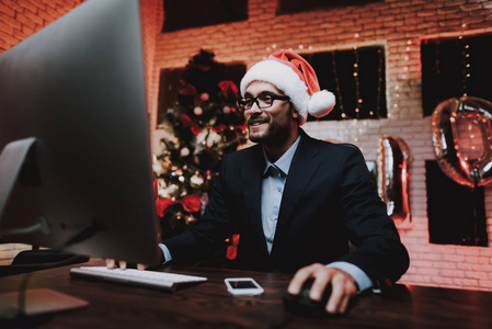 商人在除夕玩电脑。 办公室里的圣诞树。 笔记本电脑在桌子上。 商业概念。 穿黑色西装的人。 庆祝新年。 使用数字设备。 红色帽子