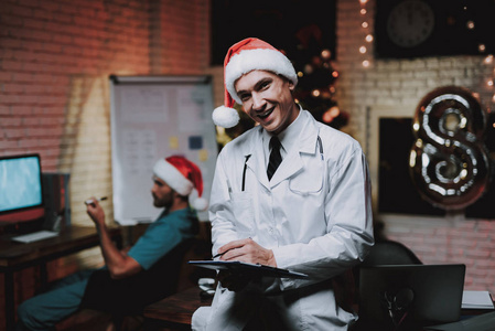 微笑的医生戴着红帽子，办公室里有药片。 新年前夜。 办公室里的圣诞树。 电脑在桌子上。 穿制服的人。 庆祝新年。 圣诞老人帽。 