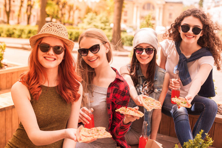 年轻的微笑女学生戴着眼镜坐在户外的长凳上，手里拿着披萨和果汁