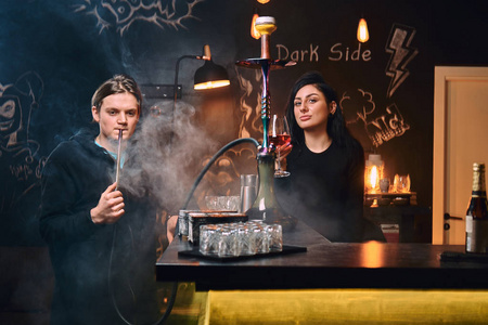英俊的家伙在连帽衫吸烟水烟和诱人的黑发妇女喝葡萄酒在夜总会或酒吧
