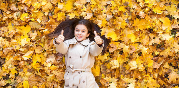 秋天的公园里，一个年轻漂亮的小女孩穿着米色外套，躺在上面的黄叶上