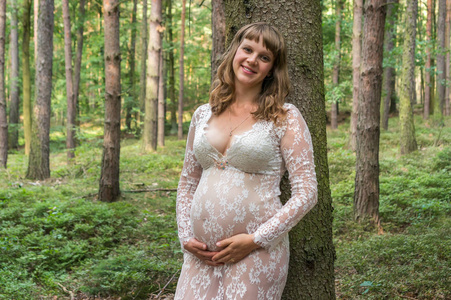 森林里穿白色蕾丝连衣裙的美丽孕妇。 怀孕概念。