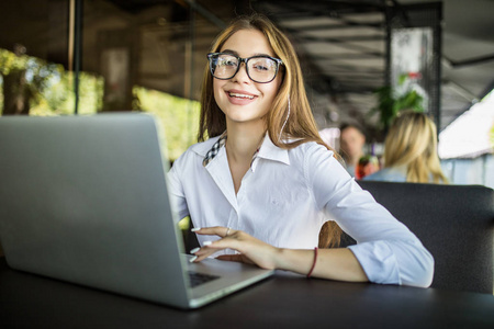 自信的年轻女性，穿着智能休闲装，戴着笔记本电脑工作的眼镜，坐在创意办公室或咖啡馆里