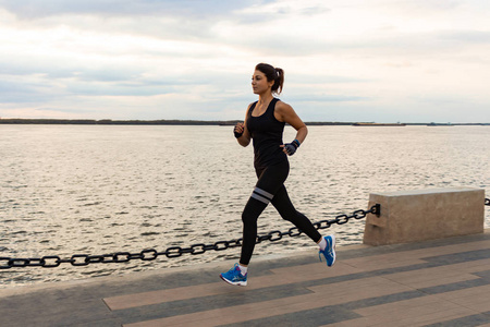 年轻而有魅力的女孩在城市公园日落时跑步。 健康的健身女人在户外跑步。 阿穆尔河在哈巴罗夫斯克地平线的背景。