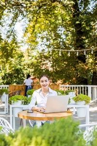 在户外工作。 戴着时髦帽子的漂亮年轻女人坐在户外时，一边拿着笔记本电脑一边微笑