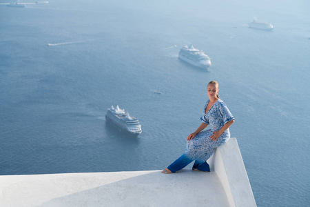 穿着白色和蓝色衣服的幸福的女人在圣托里尼, 希腊享受她的假期。从易莫洛林看火山口和爱琴海